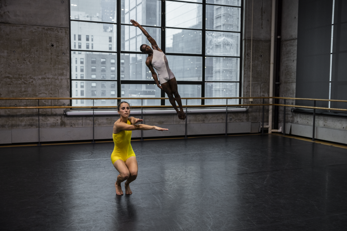 Downstage, Mariah Anton, dressed in a yellow shorty-tard, squats with her legs together, her arms extended forward in an X. Behind her, Cemiyon Barber leaps high into the air, his body a diagonal line with one arm extended over his head.